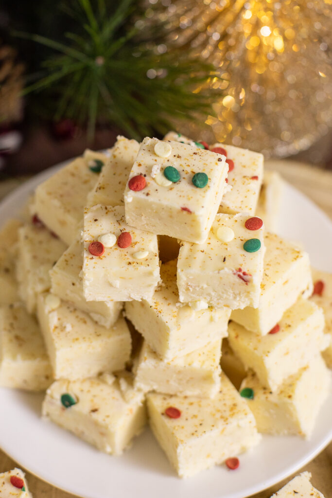 An overhead view of eggnog fudge stacked on a small white plate next to Christmas lights and an evergreen branch.