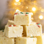 A close up of eggnog fudge pieces stacked in a pyramid shape with three on bottom, two in the center, and one on top. All pieces of fudge are topped with round red and green sprinkles, and Christmas lights are lit in the background.