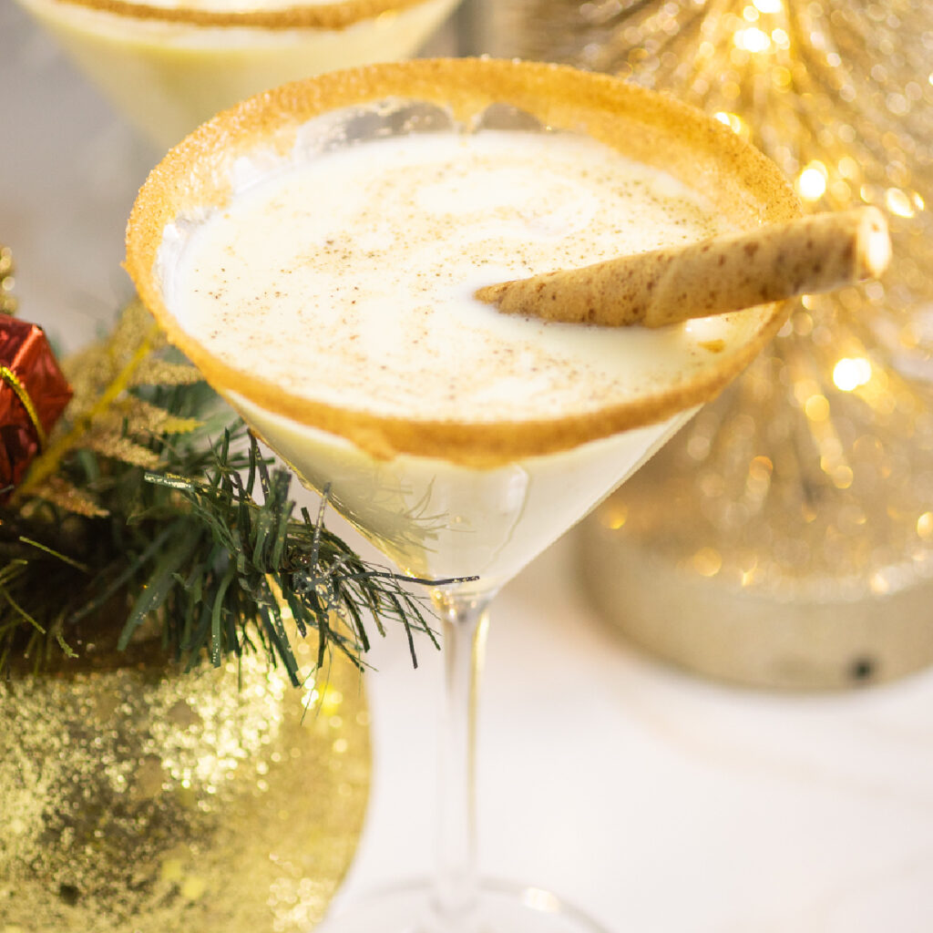 Eggnog martini in a martini glass rimmed with cinnamon sugar with a creme filled wafer cookie next to a Christmas evergreen arrangement and Christmas lights behind.