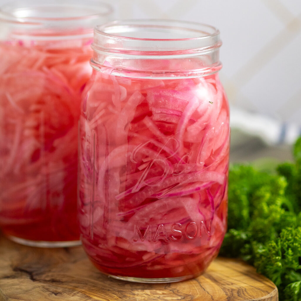 Two mason jars filled with bright pink quick pickled red onions on a small cutting board with a bunch of fresh parsley behind.