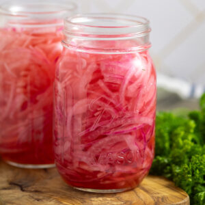 Two mason jars filled with bright pink quick pickled red onions on a small cutting board with a bunch of fresh parsley behind.