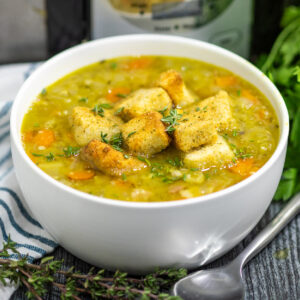 Crockpot Split Pea Soup in a white matte bowl topped with croutons and fresh thyme next to a spoon, fresh thyme sprigs, and a linen, with a slow cooker behind.