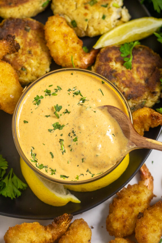 Overhead view of remoulade sauce in a small glass bowl with a wooden spoon in the center surrounded by crab cakes, fried shrimp, fresh parsley, and lemon wedges.