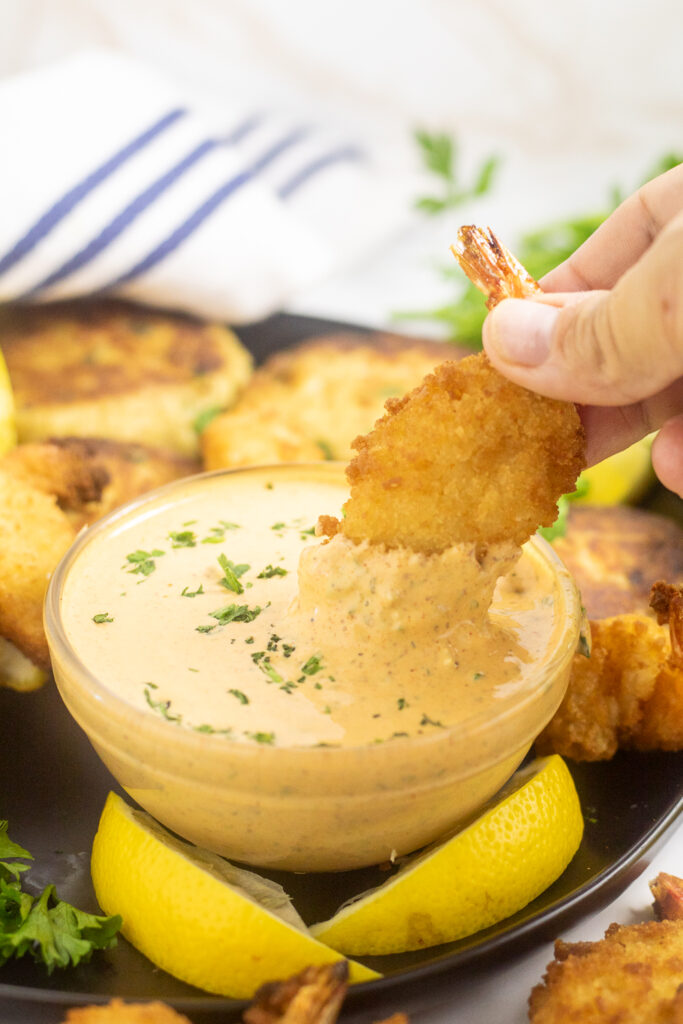 A fried shrimp being dipped into a small glass bowl of remoulade sauce with lemon wedges in front of it.