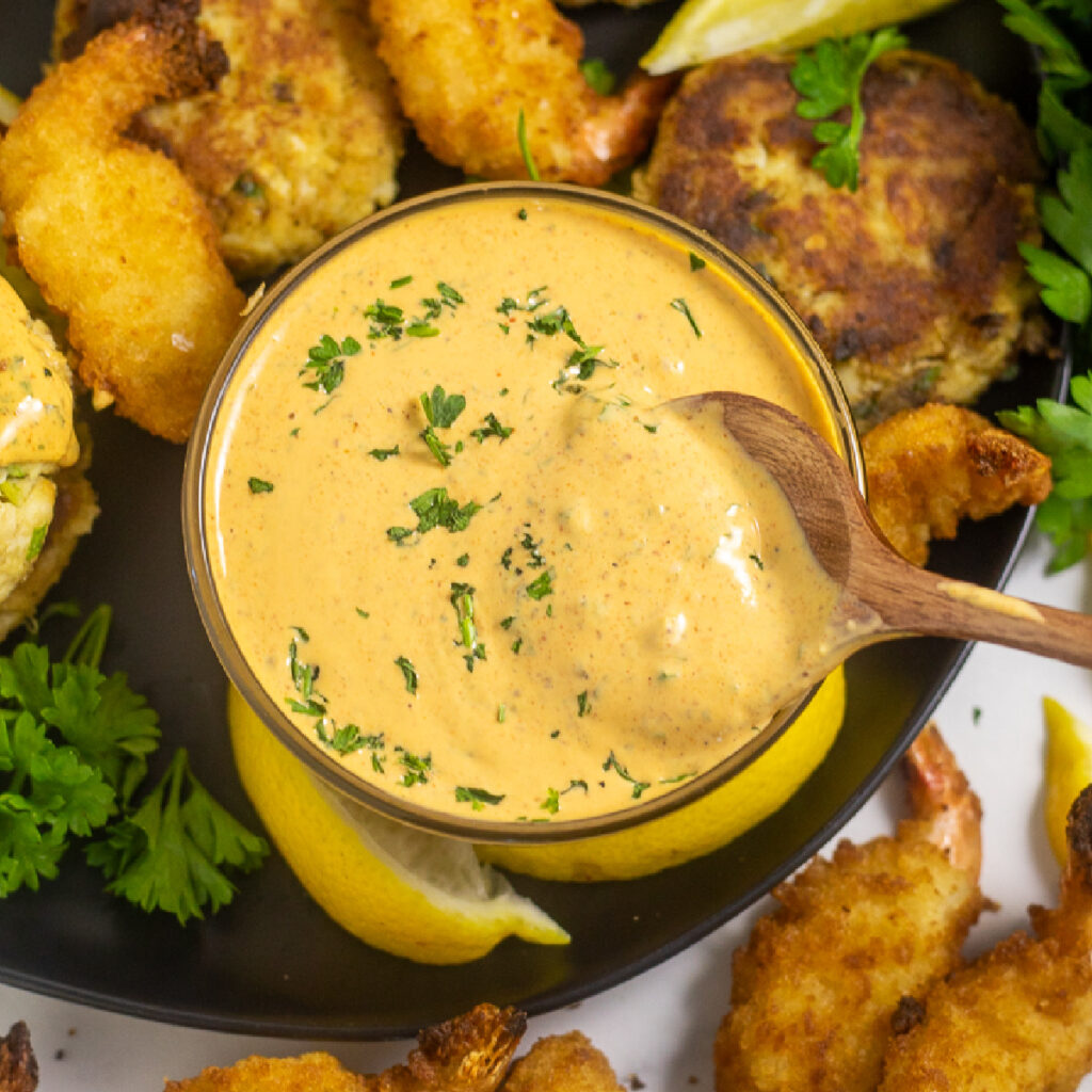 Overhead view of remoulade sauce in a small glass bowl with a wooden spoon in the center surrounded by crab cakes, fried shrimp, fresh parsley, and lemon wedges.