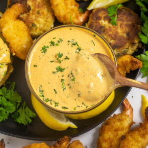 Overhead view of remoulade sauce in a small glass bowl with a wooden spoon in the center surrounded by crab cakes, fried shrimp, fresh parsley, and lemon wedges.