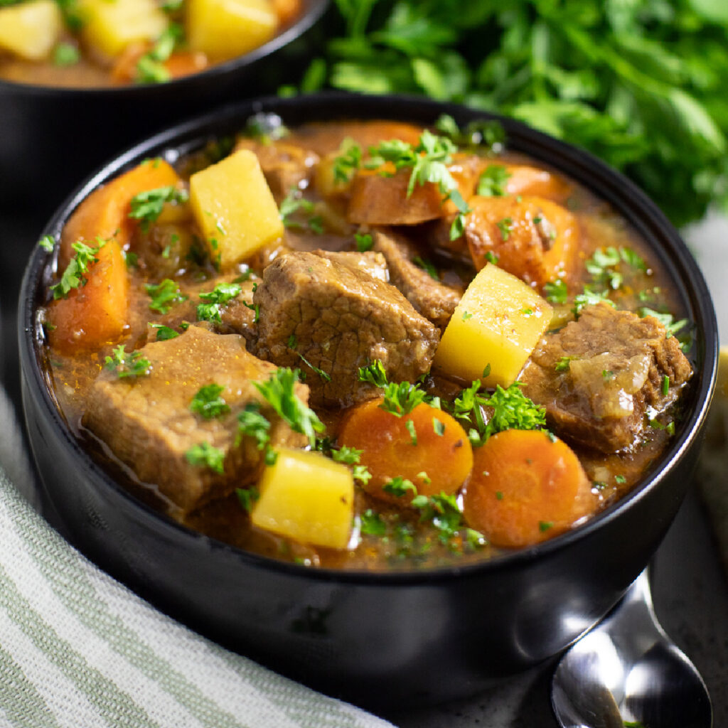 Black bowl full of Slow cooker Irish Beef stew topped with fresh chopped parsley next to a light green striped linen napkin with a second bowl of stew and a bunch of fresh parsley behind.