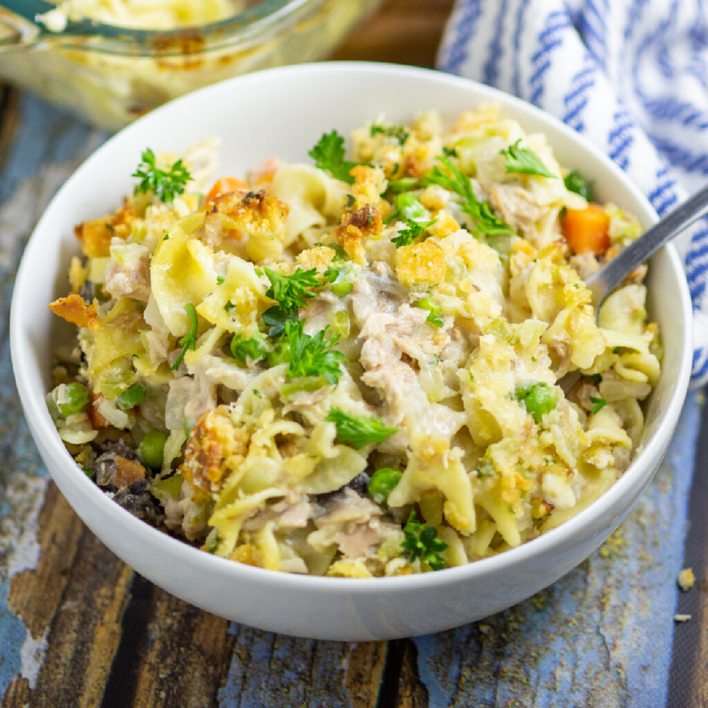 Tuna Noodle Casserole in a white bowl with a fork sticking out on a rustic wood background with a linen napkin and the full glass casserole dish behind.