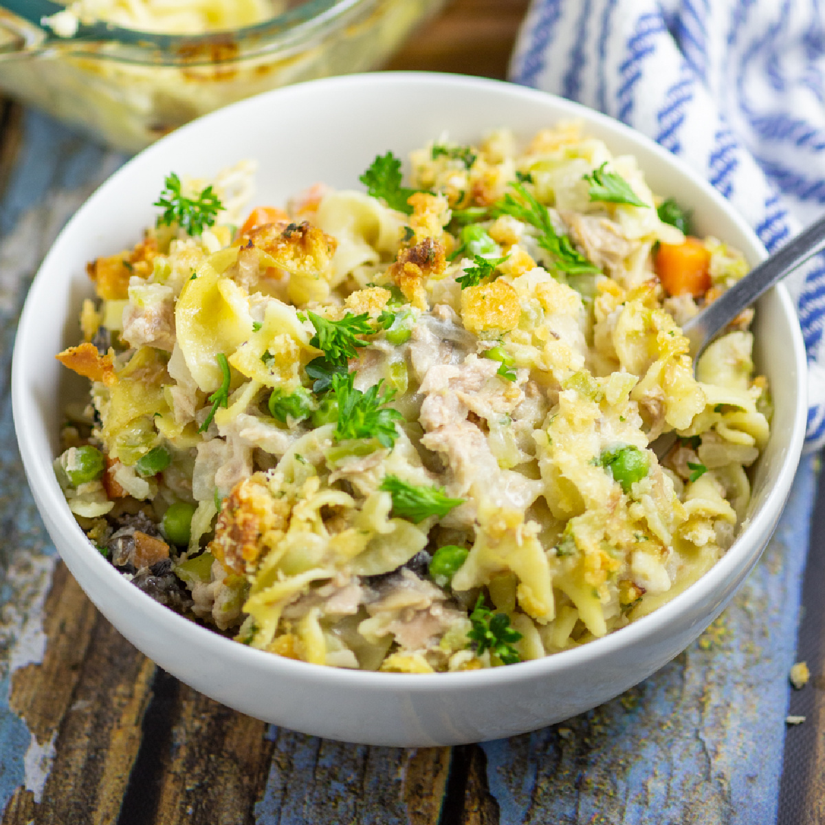 Tuna Noodle Casserole in a white bowl with a fork sticking out on a rustic wood background with a linen napkin and the full glass casserole dish behind.