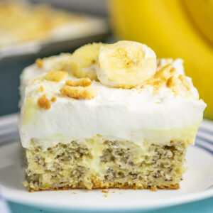 A piece of banana pudding poke cake topped with whipped cream, vanilla wafer crumbs, and fresh banana slices showing the pudding in the center of the cake, on a small white plate with bananas and the remaining cake behind.