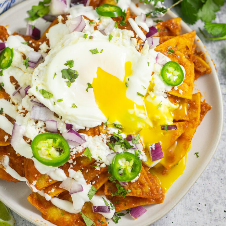 Overhead view of chilaquiles rojos topped with crema, jalapeno slices, red onion, and cilantro with a fried egg on top and the yolk running down.