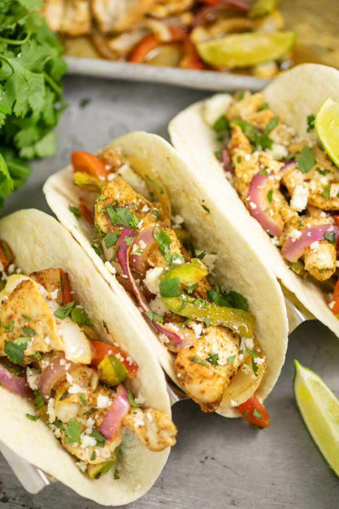 Close up of a chicken fajita in a flour tortilla topped with cotija cheese and fresh cilantro with the full sheet pan behind.