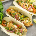 Overhead view of three chicken fajitas topped with fresh cilantro and cotija cheese in flour tortillas, next to a bunch of fresh cilantro with a sheet pan behind.
