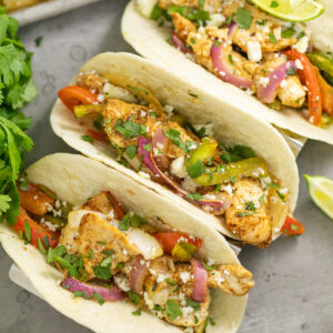 Overhead view of three chicken fajitas topped with fresh cilantro and cotija cheese in flour tortillas, next to a bunch of fresh cilantro with a sheet pan behind.