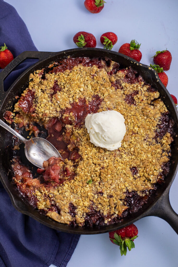 Strawberry rhubarb crisp in a cast iron skillet with a scoop missing, topped with vanilla ice cream and surrounded by fresh strawberries and a navy blue linen.