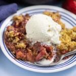 Strawberry rhubarb crisp on a small white plate topped with vanilla ice cream with a spoon in it with a navy linen and fresh strawberries behind.
