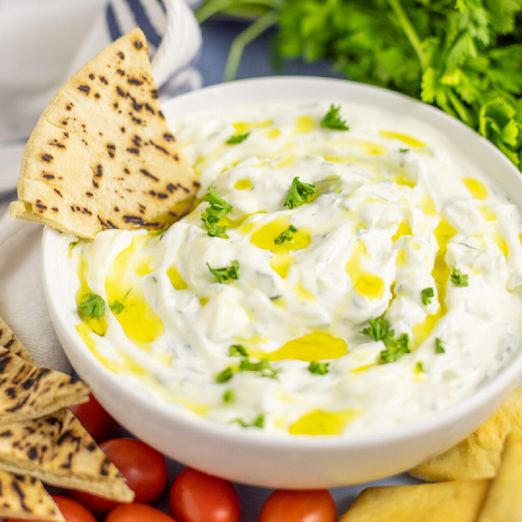 Shallow white bowl filled with homemade tzatziki topped with a drizzle of olive oil and fresh parsley with a pita wedge in the bowl. There are fresh cherry tomatoes, a bunch of parsley, pita chips, and a linen napkin next to the bowl.