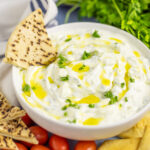 Shallow white bowl filled with homemade tzatziki topped with a drizzle of olive oil and fresh parsley with a pita wedge in the bowl. There are fresh cherry tomatoes, a bunch of parsley, pita chips, and a linen napkin next to the bowl.