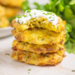A stack of four air fryer corn fritters with sour cream and chives on the top on and a bite taken out of it. There is a plate of fritters and a bunch of fresh parsley behind the stack.