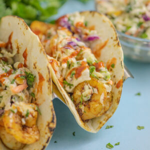 Air Fryer Shrimp Tacos topped with creamy cilantro slaw on a blue background with additional slaw and fresh cilantro in the background.