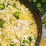A chip in melty baked elote dip in a cast iron skillet topped with cotija cheese and fresh jalapeno slices with chips, lime wedges, and fresh cilantro around the skillet on a concrete background.