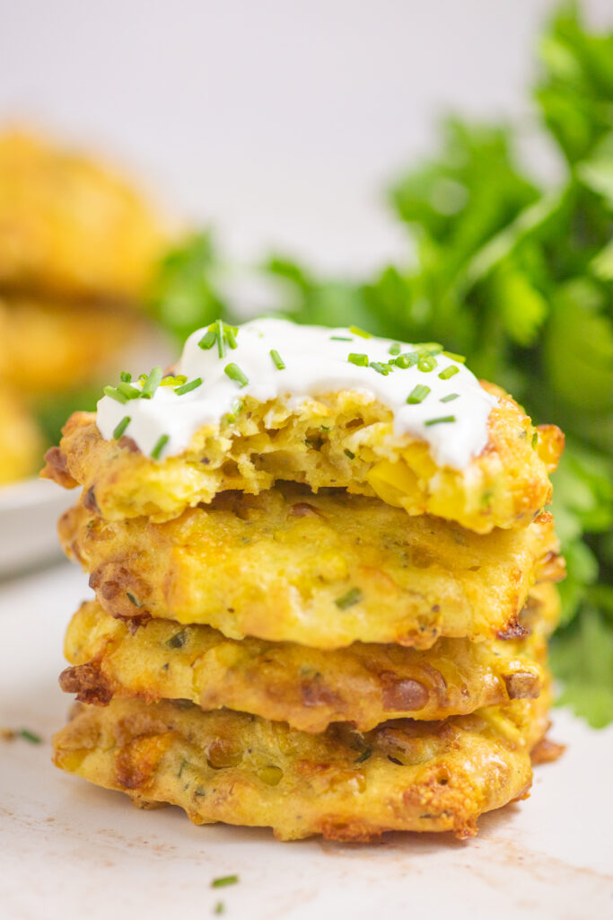 A stack of four air fryer corn fritters with sour cream and chives on the top on and a bite taken out of it. There is a plate of fritters and a bunch of fresh parsley behind the stack.