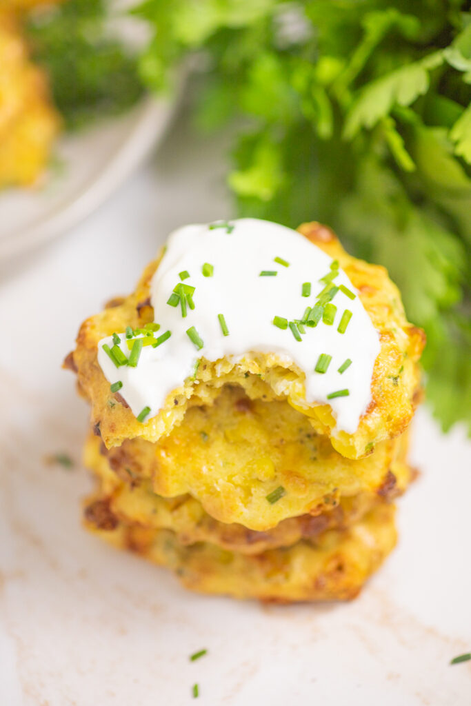Overhead view of a stack of air fryer corn fritters with sour cream and chives on top and a bite taken out of the top one. There's a bunch of fresh parsley next to the stack.