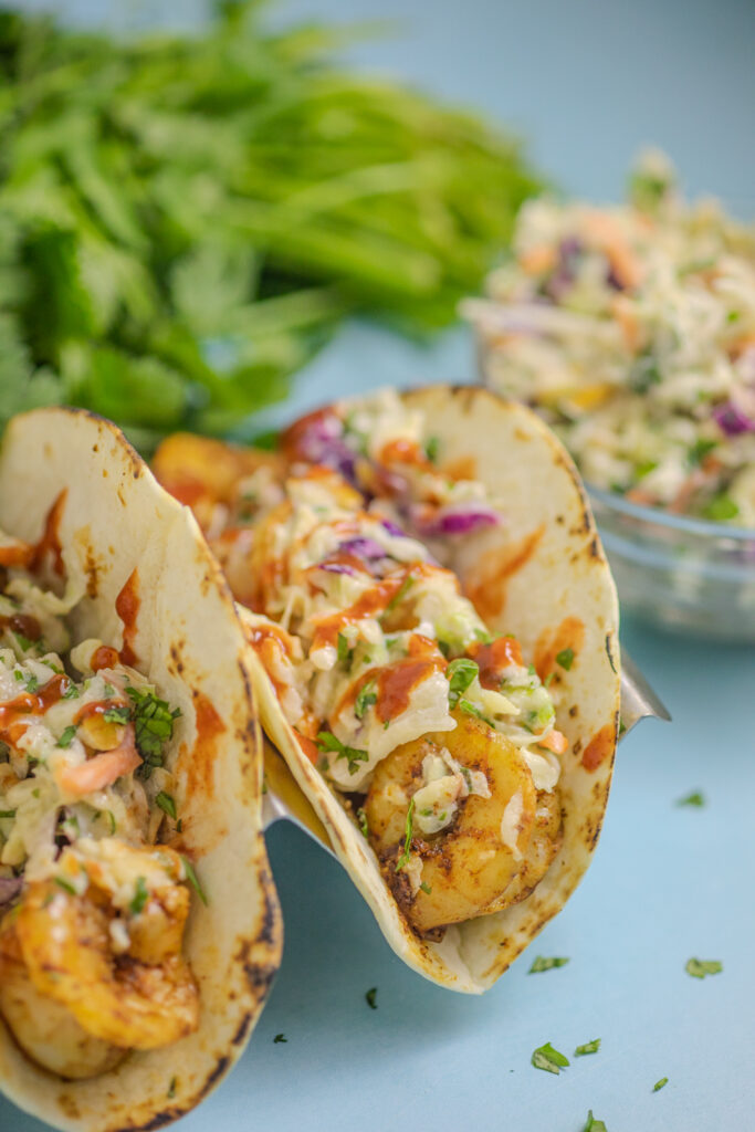 Air Fryer Shrimp Tacos topped with creamy cilantro slaw on a blue background with additional slaw and fresh cilantro in the background.