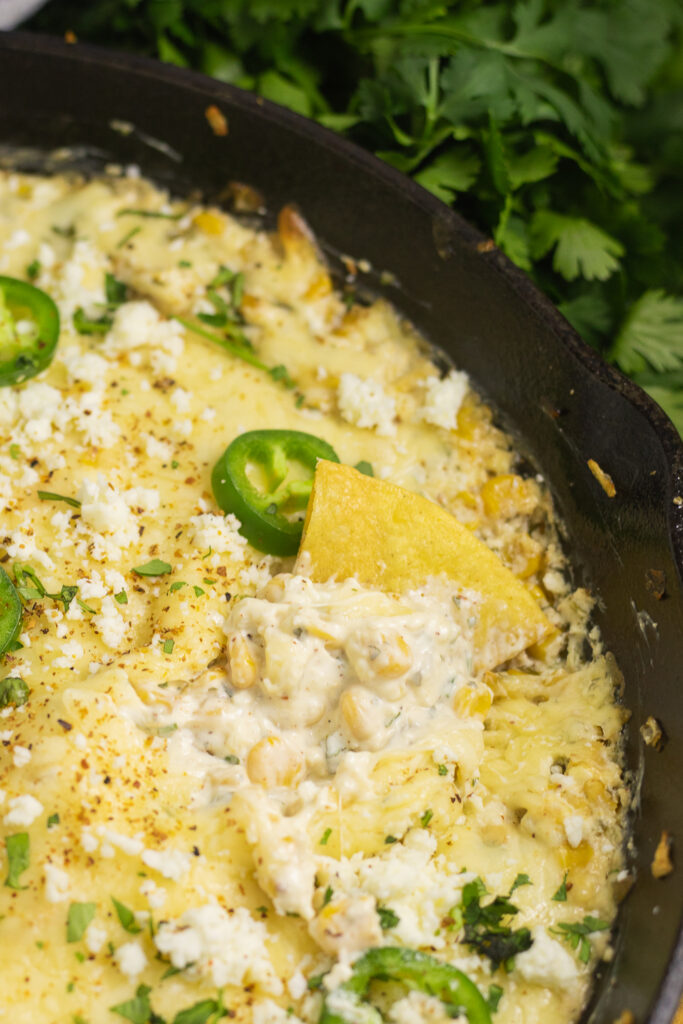 A chip in melty baked elote dip in a cast iron skillet topped with cotija cheese and fresh jalapeno slices with chips, lime wedges, and fresh cilantro around the skillet on a concrete background.