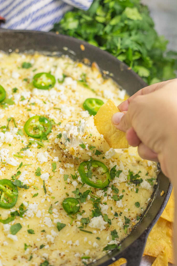 A hand with a chip scooping warm elote dip from a cast iron skillet with fresh cilantro behind.