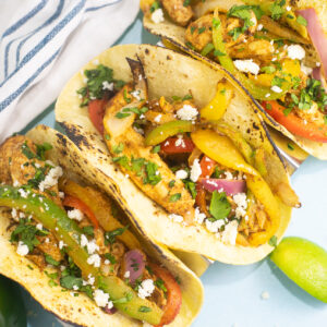 Overhead view of three air fryer chicken fajitas in corn tortillas topped with fresh cilantro on a blue background next to lime wedges and a linen napkin.