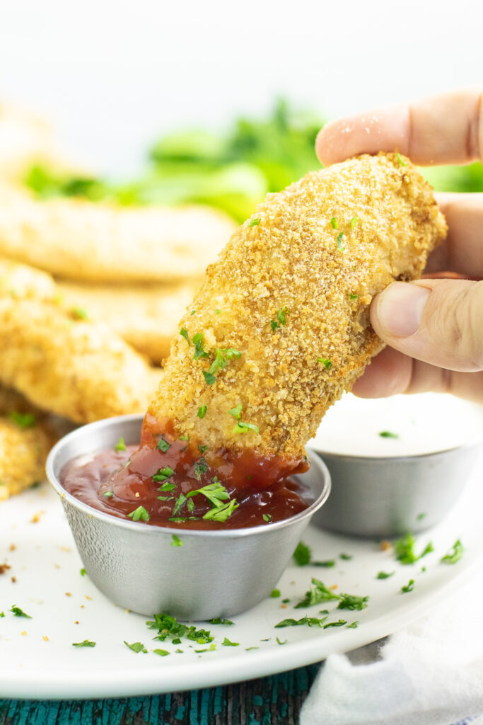 An air fryer chicken tenders being dunked into a small metal bowl of ketchup.