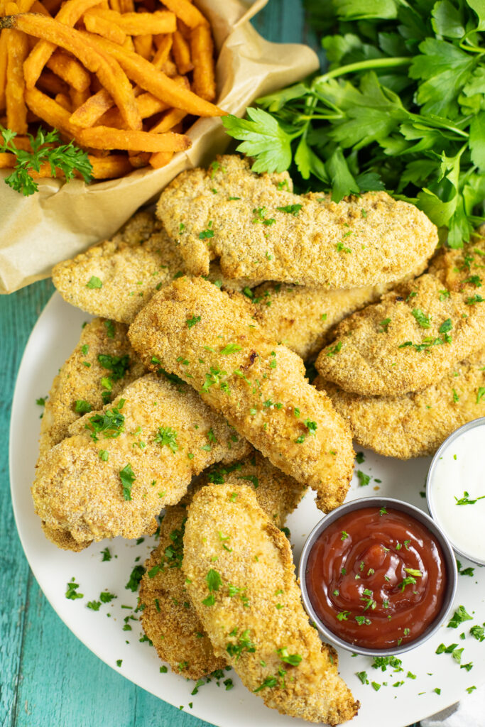 Overhead view of air fryer chicken tenders on a large white plate topped with freshly chopped parsley with a small metal bowl of ketchup and a small metal bowl of ranch dip on the plate. A bunch of fresh parsley and a basket of sweet potato fries is behind, all on a bright teal wood background.