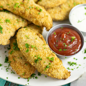 Air fryer chicken tenders stacked on a plate next to a small metal bowl of ketchup, all topped with freshly chopped parsley.