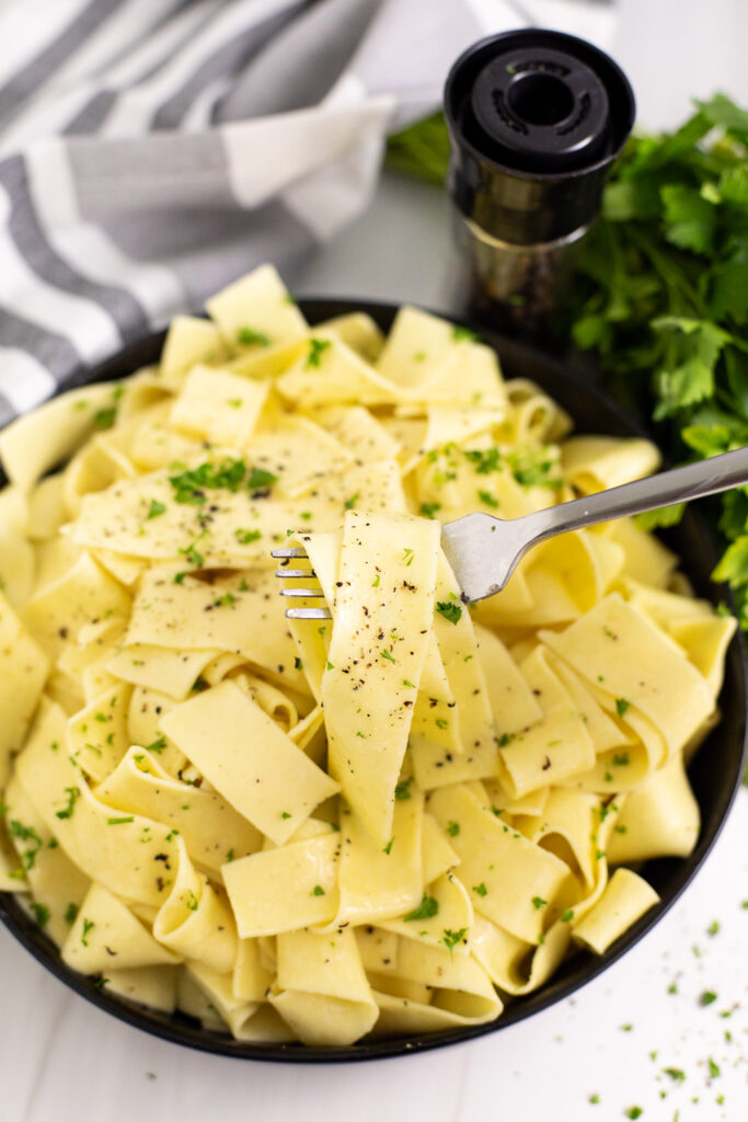 Overhead view of buttered noodles in a black bowl topped with black pepper and parsley with a fork pulling out a noodle.