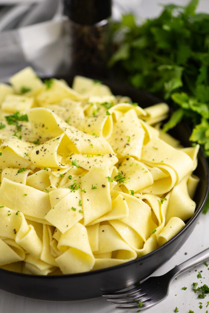 Side view of buttered noodles in a black bowl topped with black pepper and parsley with fresh parsley and a linen napkin behind it.