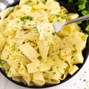 Overhead view of buttered noodles in a black bowl topped with black pepper and parsley with a fork pulling out a noodle.