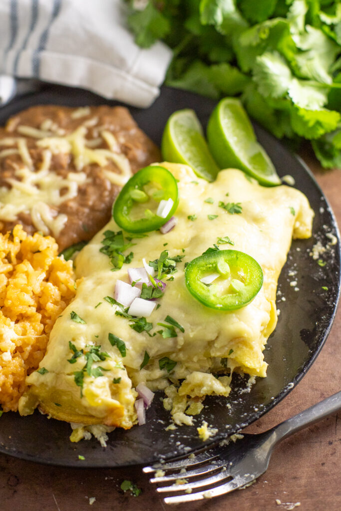 Two cheesy chicken enchiladas on a black matte plate with beans rice. One enchilada has a bite taken out of it. The plate is on a rustic background with a linen napkin and bunch of fresh cilantro behind.
