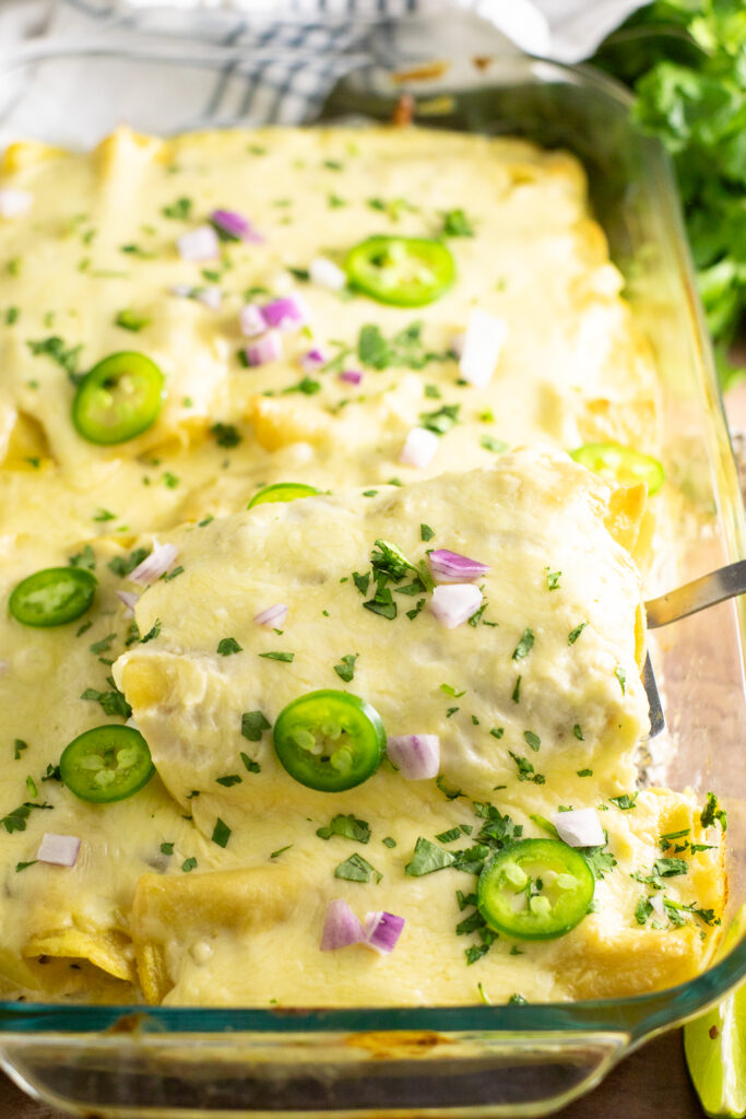 Two cheesy chicken enchiladas being lifted from a casserole dish filled with enchiladas topped with cheese sauce, fresh cilantro, jalapeno slices, and chopped red onion.