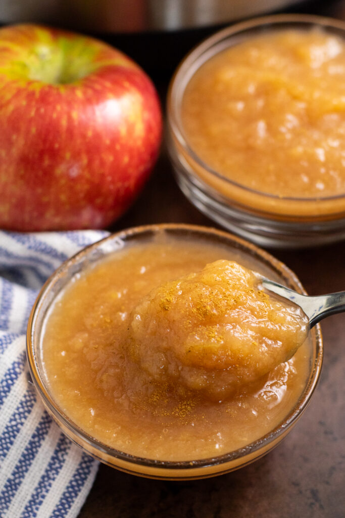 A small glass bowl filled with applesauce with a spoon taking a scoop out with a second bowl of applesauce, a red apple, and a linen napkin behind it on a rustic background.