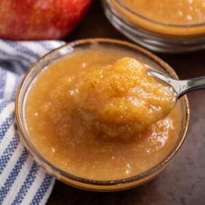 A small glass bowl filled with applesauce with a spoon taking a scoop out with a second bowl of applesauce, a red apple, and a linen napkin behind it on a rustic background.