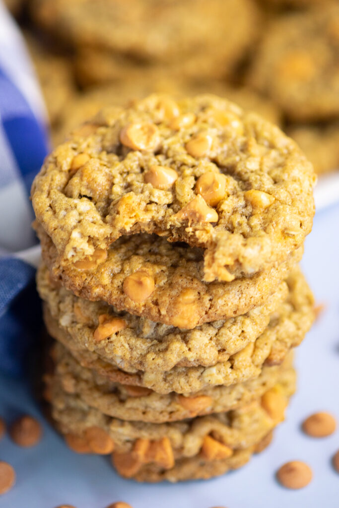 Overhead view of a stack of oatmeal scotchies with a bite taken out of the top one. The stack is surrounded by butterscotch chips and a blue checkered linen napkin.