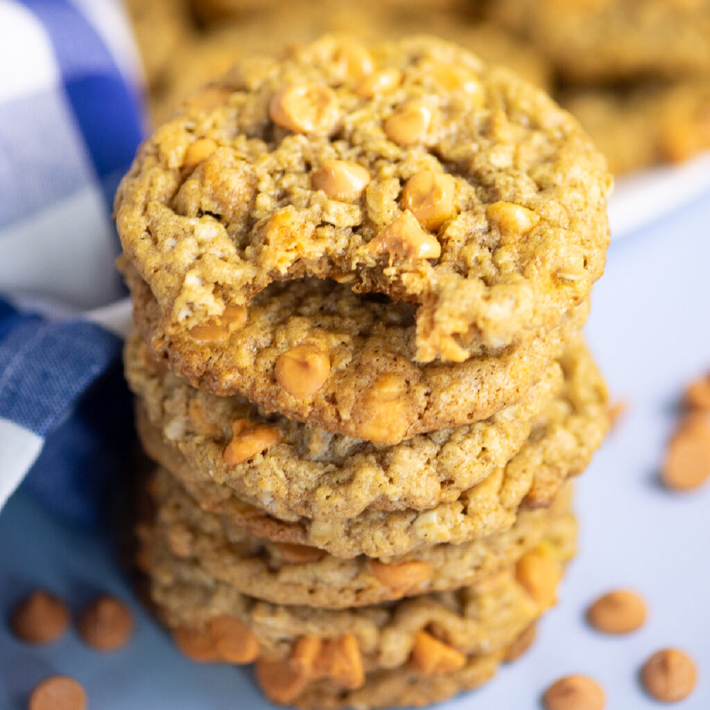 Overhead view of a stack of oatmeal scotchies with a bite taken out of the top one. The stack is surrounded by butterscotch chips and a blue checkered linen napkin.