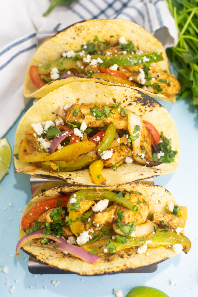 Overhead view of three air fryer chicken fajitas in corn tortillas topped with fresh cilantro on a blue background next to lime wedges and a linen napkin.