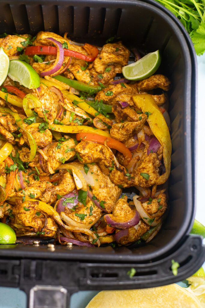 Fajita-seasoned chicken with peppers and onions in an air fryer basket topped with fresh cilantro and lime wedges.