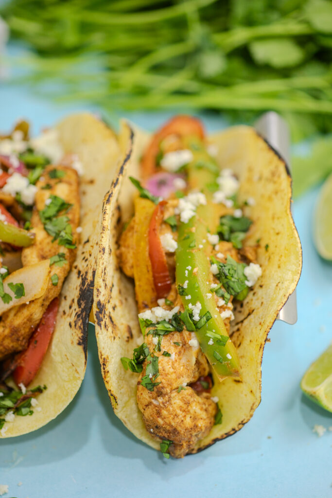 Close up of an air fryer chicken fajita topped with cotija cheese and fresh cilantro with a bunch of fresh cilantro behind on a blue background.