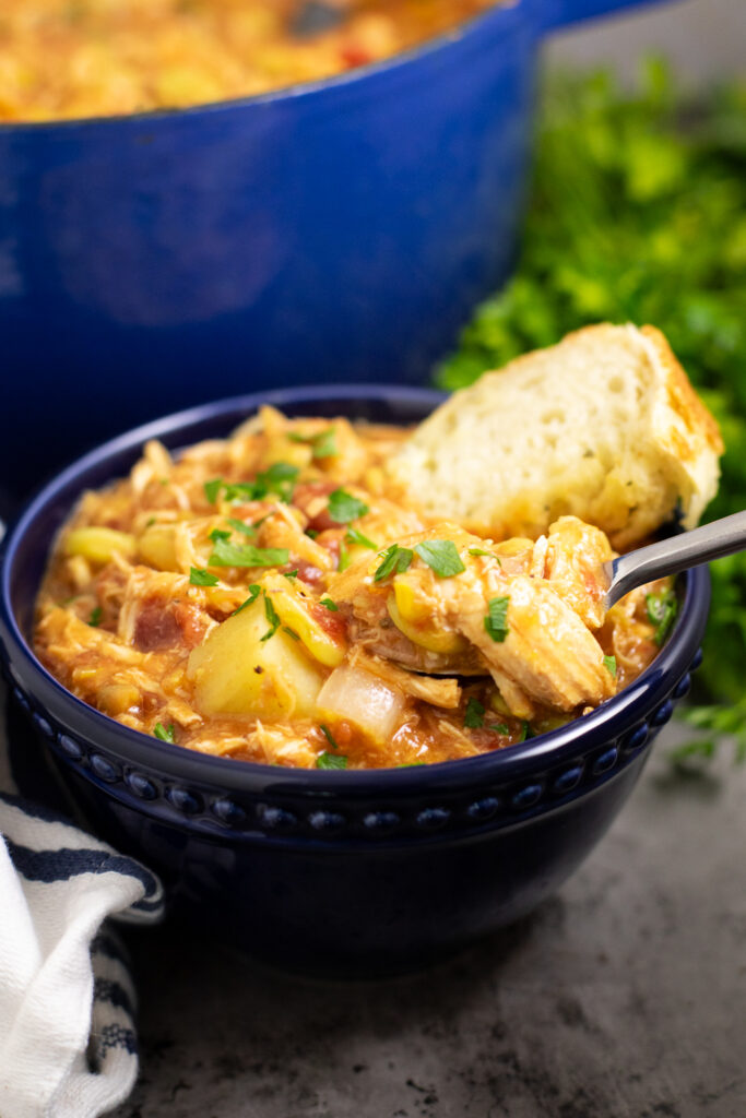 Brunswick stew in a dark blue bowl topped with fresh parsley. A piece of crusty bread is in the stew and a spoon is taking a bite out.