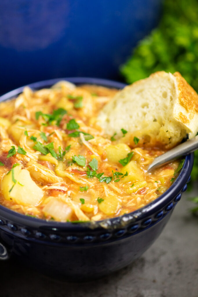 Brunswick stew in a dark blue bowl topped with fresh parsley. A spoon and a piece of crusty bread of in the stew with a