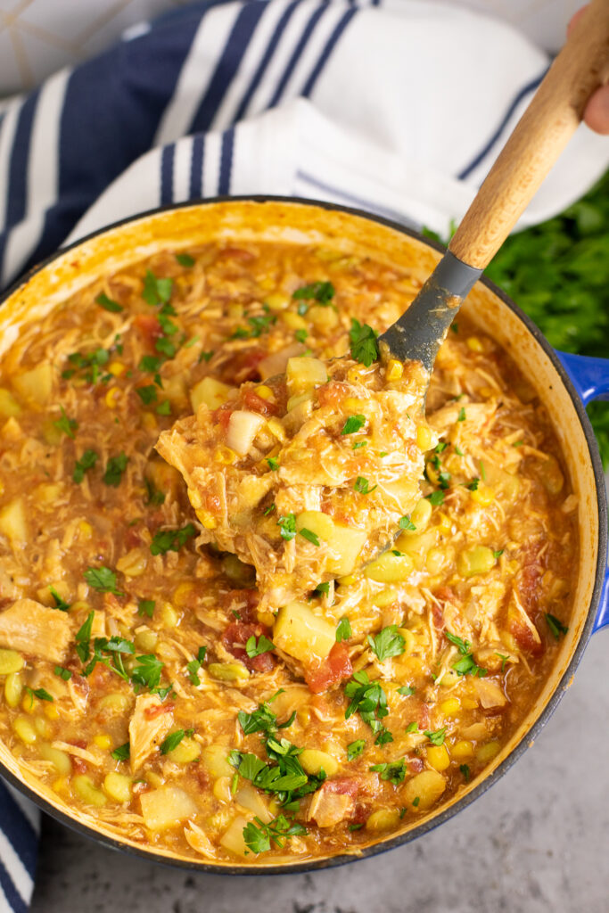 Overhead view of Brunswick stew in a large Dutch oven with a ladle scooping some out. The pot is surrounded by a bunch of fresh parsley and a linen napkin.