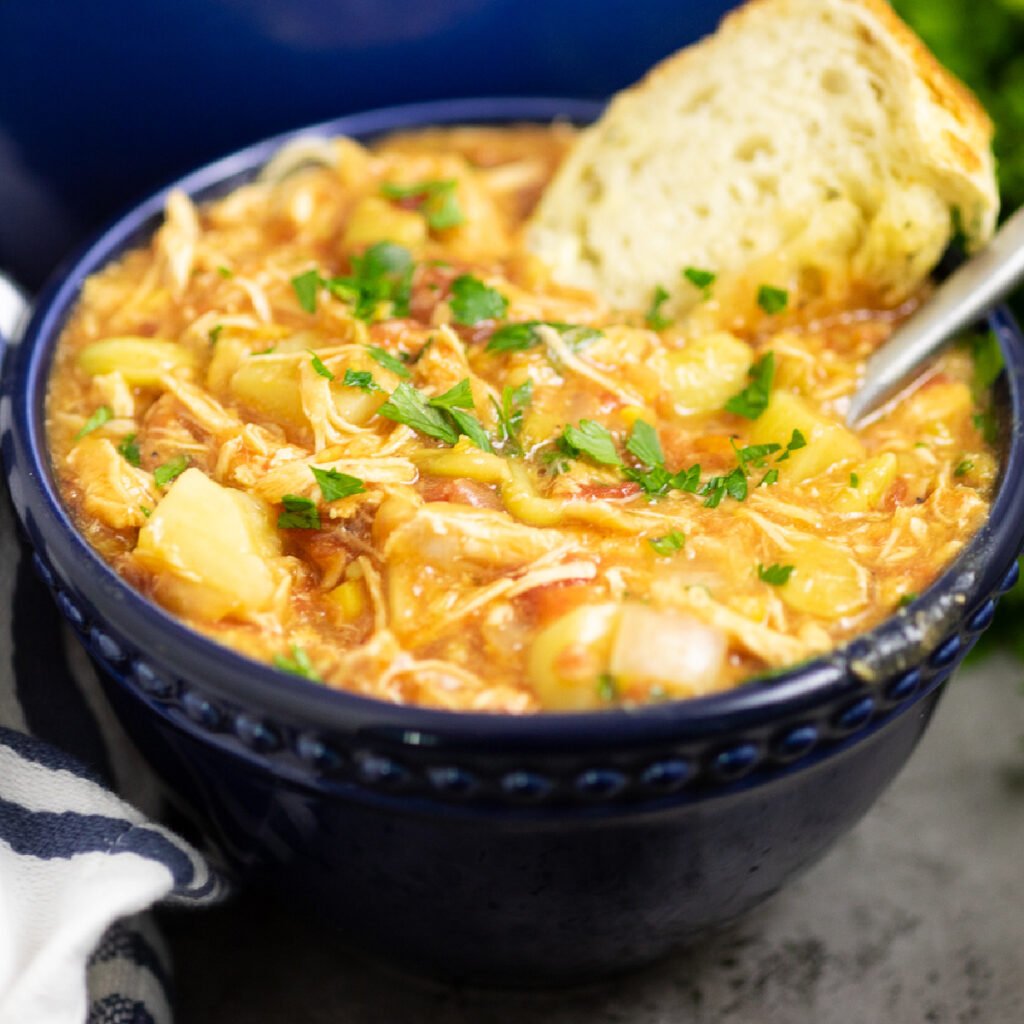 Brunswick stew in a dark blue bowl topped with fresh parsley. A spoon and a piece of crusty bread of in the stew.
